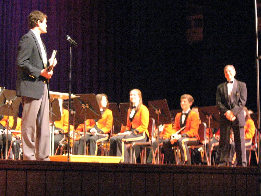 Eine Gruppe von Kindern sitzt auf Stühlen und schaut auf einen Mann in einem schwarzen Anzug, der in der Nähe eines Mikrofons steht, mit einem großen Vorhang dahinter.