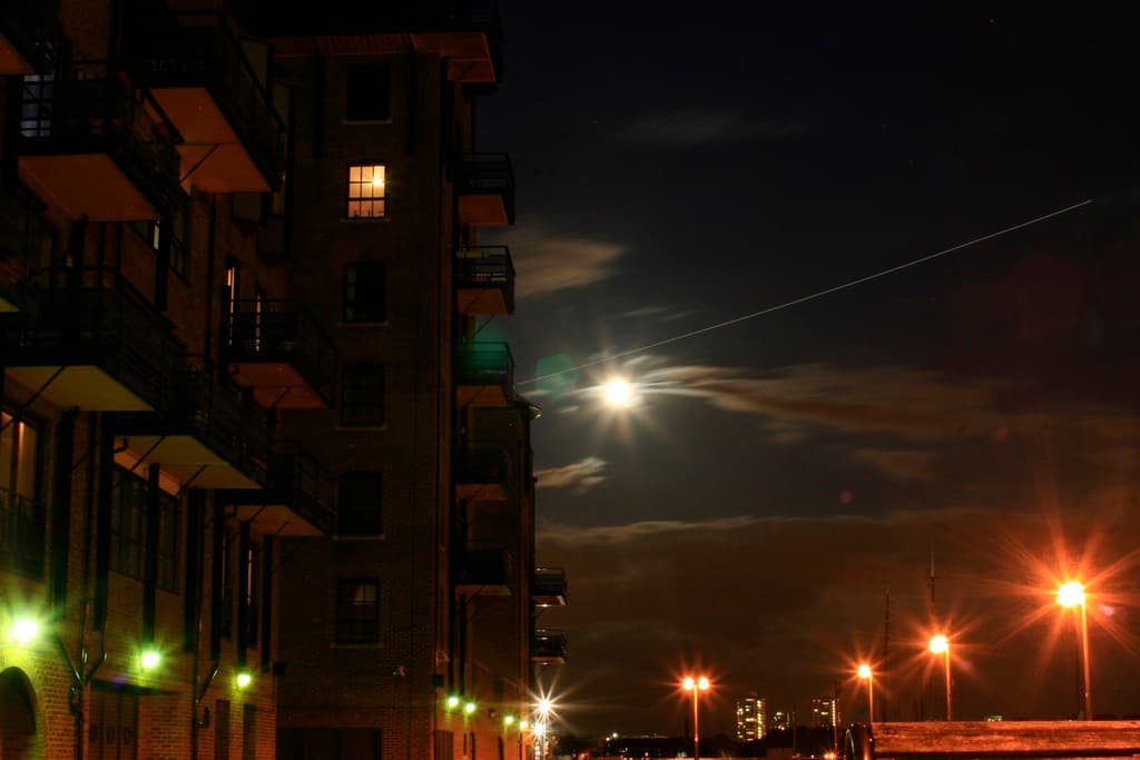Eine nächtliche Stadtansicht mit einem prominenten Gebäude mit zahlreichen Fenstern und Balkonen sowie mehreren Straßenlaternen auf der rechten Seite.