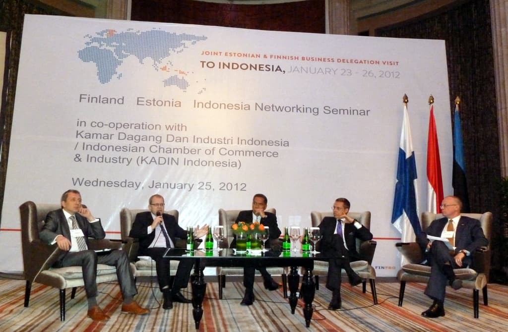 Eine Konferenzbühne mit Menschen, Stühlen, einem Mikrofon, Kabeln, einem Tisch, Flaschen, Blumen, Gläsern und anderen Gegenständen, mit Flaggen und einem Banner in der Mitte und einem Gebäude mit Vorhängen im Hintergrund.