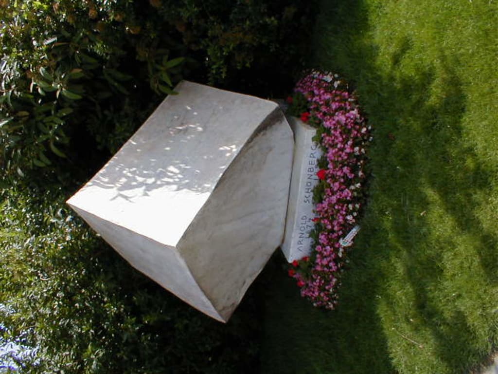 Ein Denkmal mit roten und pinken Blumen davor, umgeben von vielen Bäumen im Hintergrund.