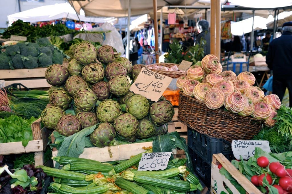 Körbe voller Obst, Gemüse und kleine Schilder mit Text, mit Zelten und wenigen Menschen im Hintergrund.