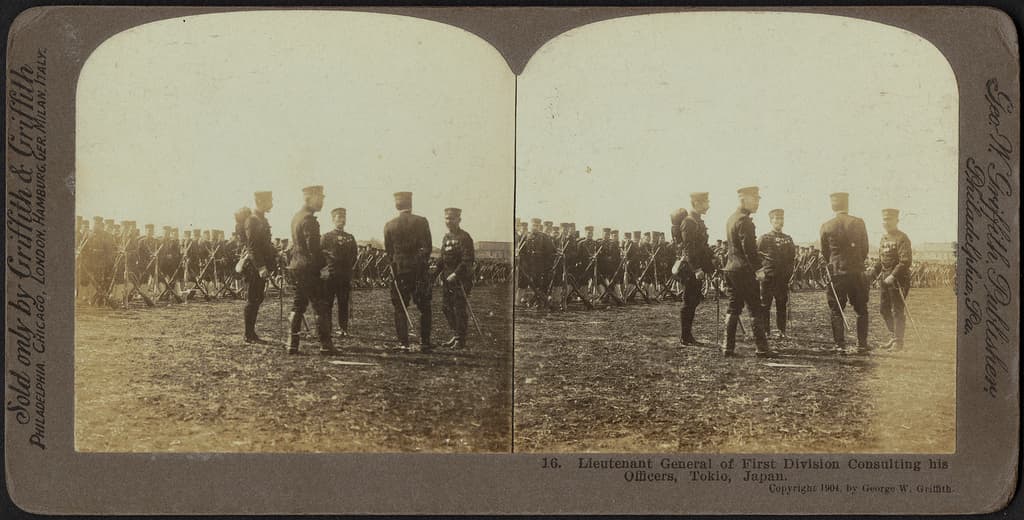 Ein altes Gruppenfoto der Militärpersonen, die vorne stehen, mit Soldaten im Hintergrund, die Gewehre halten.