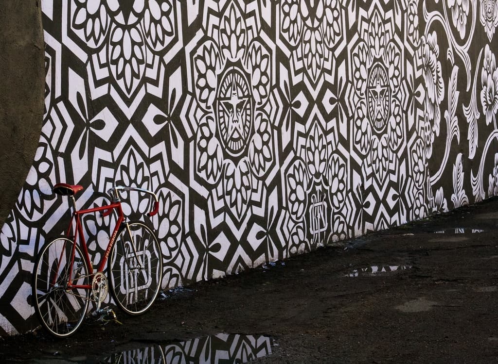 Eine Straße mit einem geparkten Fahrrad auf der linken Seite, einer Wand mit Gemälden in der Mitte und einer Außenkulisse.