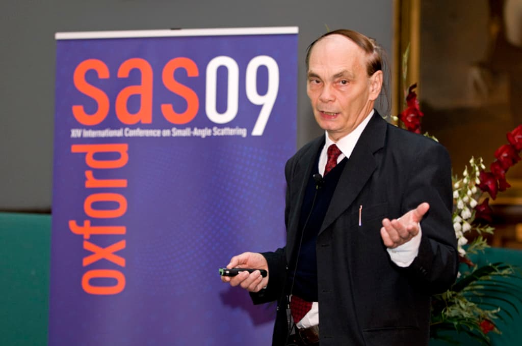 Ein älterer Mensch in einem Anzug hält ein Objekt, während er spricht, mit einem Banner, einer Wand, einem Foto Rahmen und Blumen im Hintergrund.