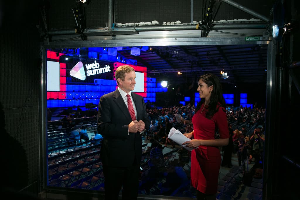 Ein Mann im Anzug und eine Frau in einem roten Kleid halten Papiere und stehen zusammen, lächeln und sind von einer Gruppe von Menschen und Kameras umgeben, und es sind Text und Lichter zu sehen.