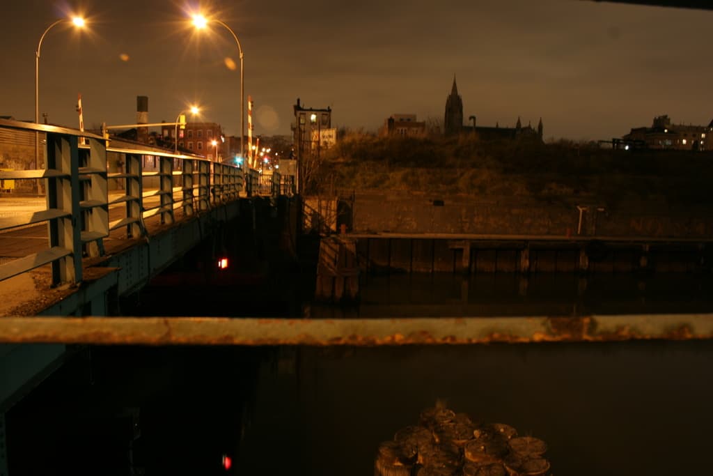 Eine Brücke spannt einen Fluss mit mehreren Straßenlaternen auf ihrer Länge und zahlreichen Gebäuden im Hintergrund.