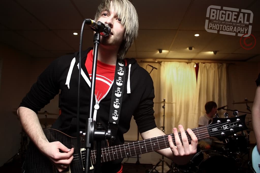Ein schwarzes Mikrofon auf einem StÃ¤nder im Vordergrund, Musiker mit Instrumenten in der Mitte, VorhÃ¤nge im Hintergrund, Scheinwerfer Ã¼ber ihnen und sichtbarer Text oben rechts.