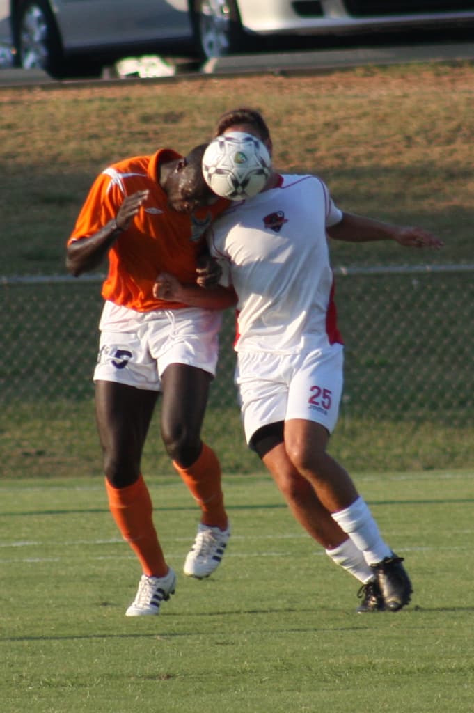 Zwei Spieler in einem eingezäunten Bereich, einer in einem orangen T-Shirt, der den Ball mit dem Kopf schlägt, mit Autos und Gras im Hintergrund.