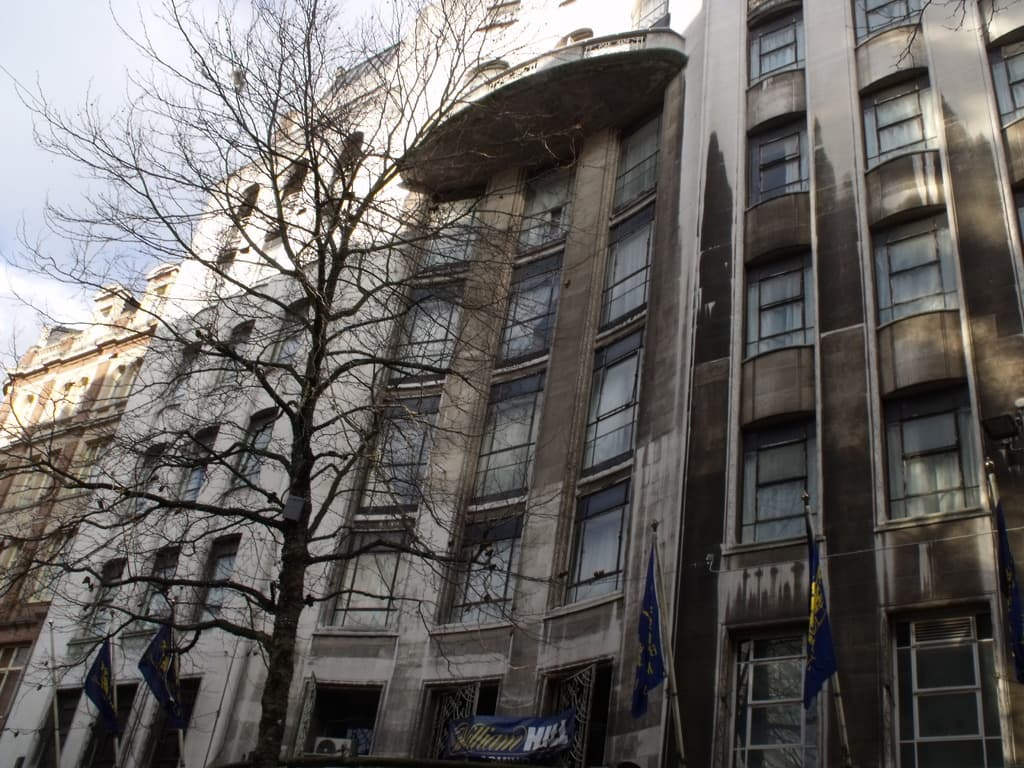 Geb√§ude mit Fenstern, einer Fahne und Flaggen an Masten unten sowie einem Baum mit √Ąsten.