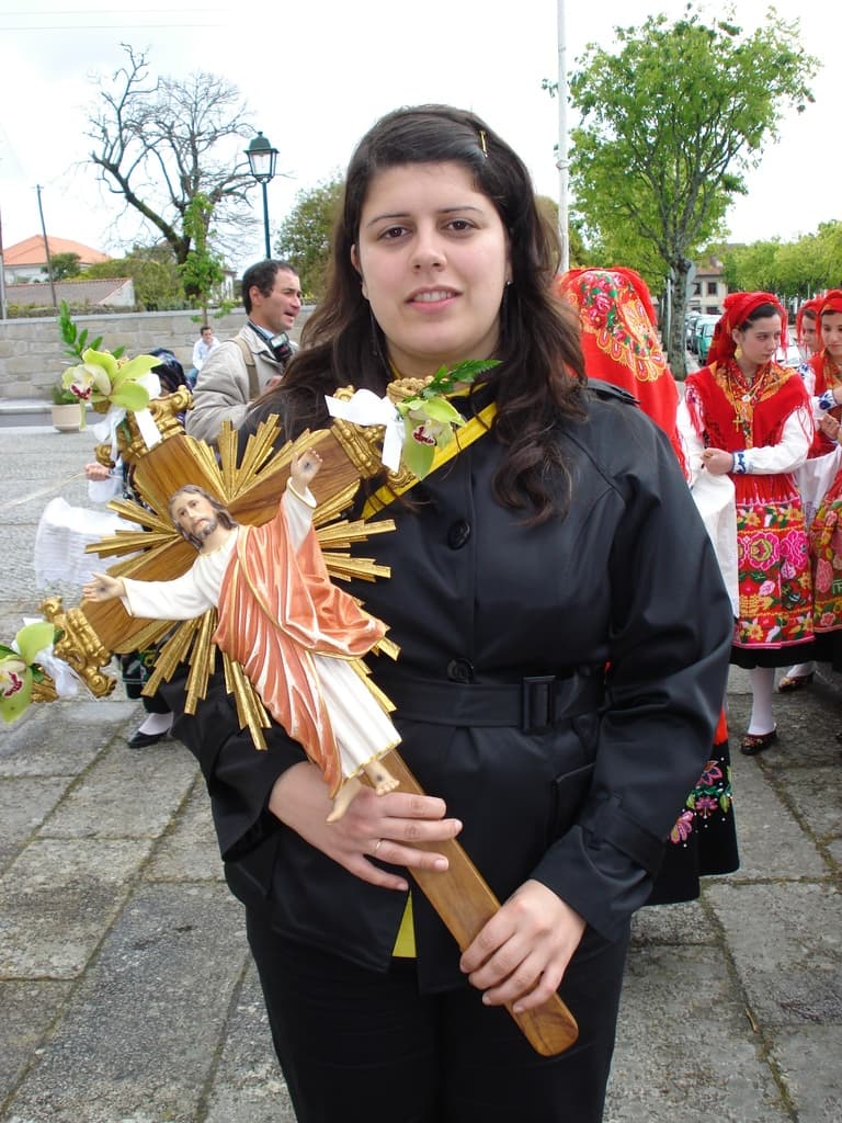 Eine Frau hält ein Abbild von Jesus Christus in der Hand, mit ein paar anderen Menschen, die dahinter auf dem Bürgersteig stehen, und Bäumen, Laternenpfählen, Pflanzen, Straßen, Gebäuden und einem Metallpfahl im Hintergrund.