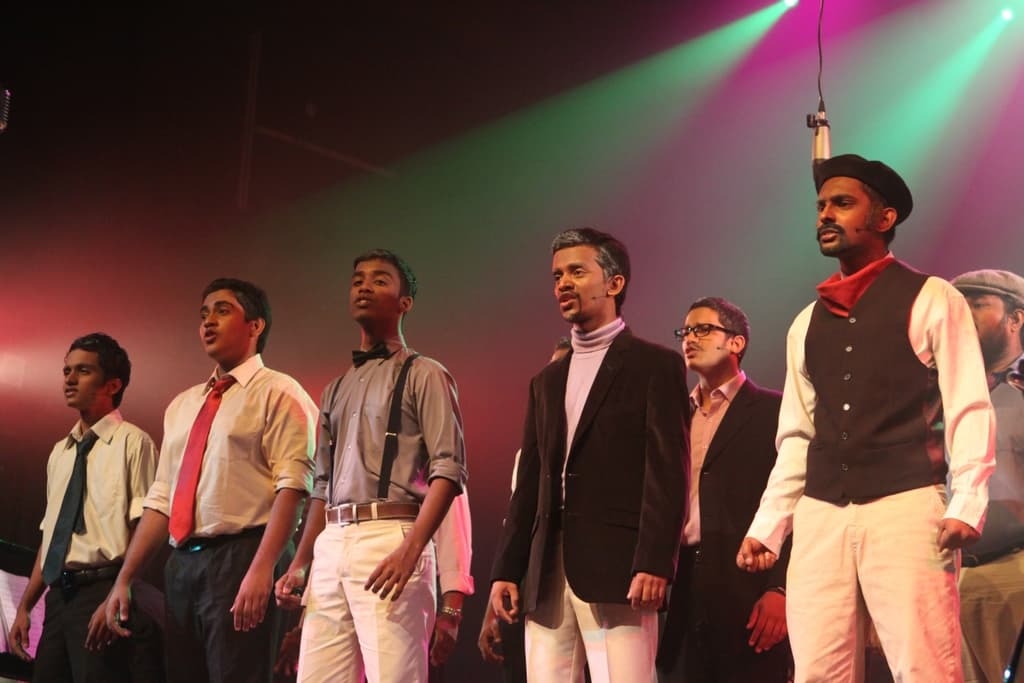 Eine Gruppe von Männern steht auf einer Bühne und spielt ein Drama, während grüne Scheinwerfer hinter ihnen leuchten.