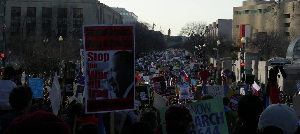 Eine Gruppe von Menschen demonstriert auf einer Straße, hält Plakate, mit Gebäuden, Bäumen und Laternen im Hintergrund unter einem klaren Himmel.