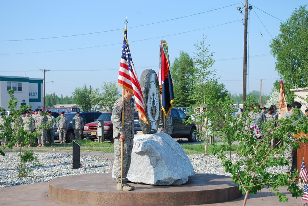 Zwei Soldaten stehen mit Fahnen auf beiden Seiten eines Gedenksteins, dahinter sind Pflanzen, Autos, weitere Soldaten, Strommasten mit Kabeln und Gebäude zu sehen.