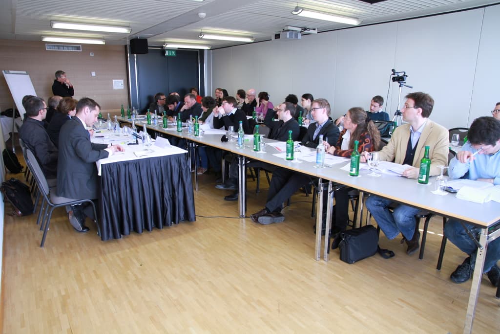 Ein Büroraum mit Mitarbeitern an Tischen, Rucksäcke auf dem Boden, ein Mann im Hintergrund und verschiedene Gegenstände wie Flaschen, Papiere, eine Kamera, Lautsprecher, Schilder, Projektor und Lampen.