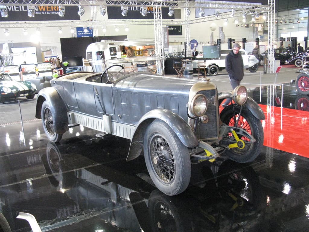 Ein Auto ist in der Mitte einer Ausstellungshalle zu sehen, umgeben von einem Zaun, mit zahlreichen anderen Autos im Hintergrund und auf der rechten Seite eine Person, Lichter und Werbetafeln oben.
