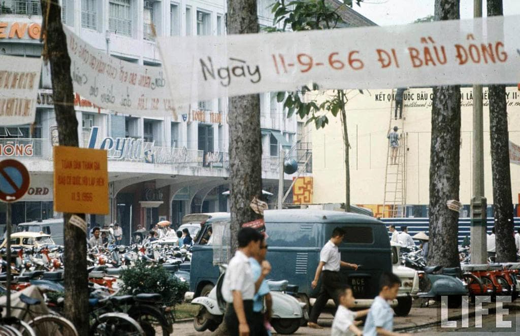 Eine Straßenszene mit Menschen, Fahrrädern, einem Lieferwagen, Autos im Vordergrund, Gebäuden und Bäumen im Hintergrund sowie verschiedenen Bannern, Tafeln und einem Schild auf der linken Seite.