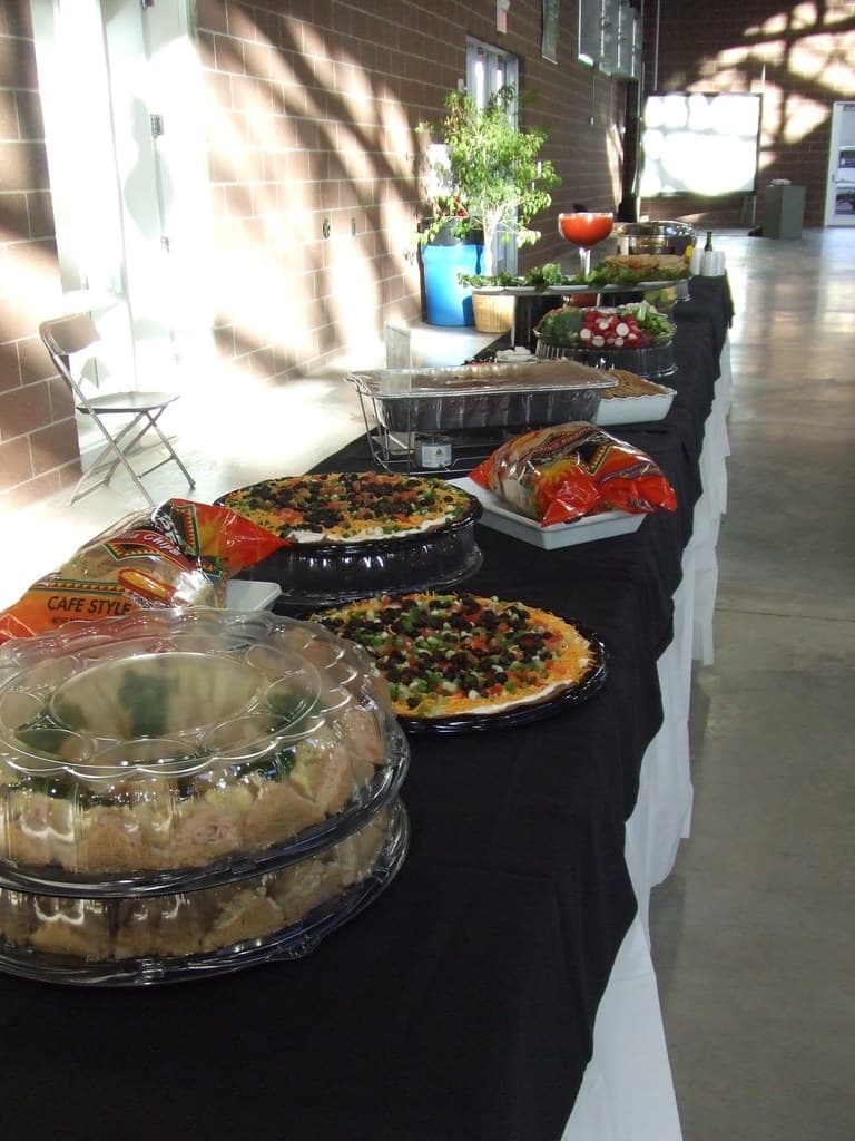 Ein Tisch mit einer schwarzen und weißen Decke darauf, auf dem ein Teller mit Pizza, ein Tablett mit Plastiktüten und Gefäße mit Essen stehen; daneben ein Stuhl, mit einem braunen Gebäude, einem Blumentopf und einem blauen Müllcontainer im Hintergrund.
