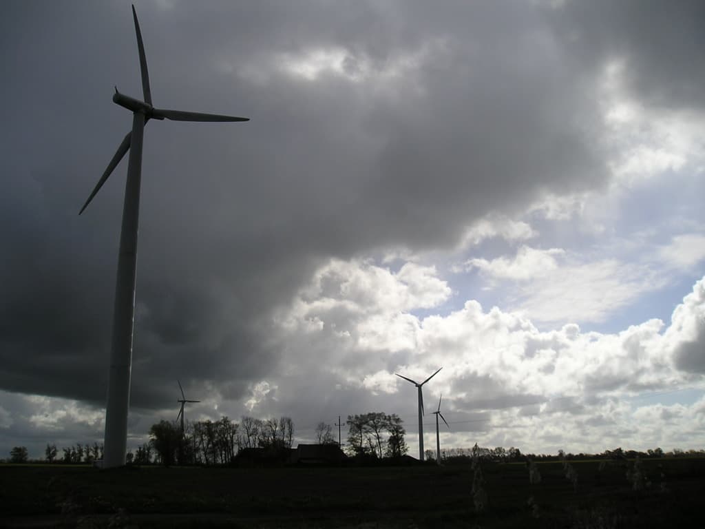 Windräder mit Bäumen mit Zweigen und Blättern im Vordergrund, Wolken am Himmel und ein Haus im Hintergrund.