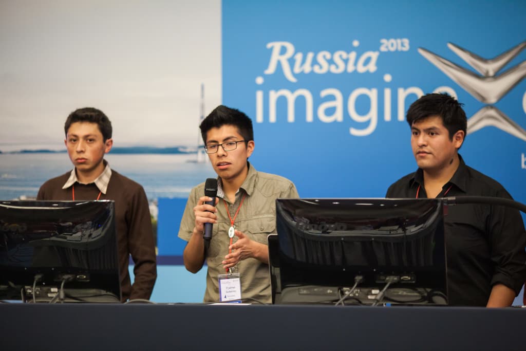 Drei Jungen stehen neben einem Schreibtisch mit zwei Computersystemen, einer spricht in ein Mikrofon und trägt eine ID-Karte mit rotem Tag, vor einer Wand mit einem Werbeplakat, auf dem "Russia Imagine 2013" steht.