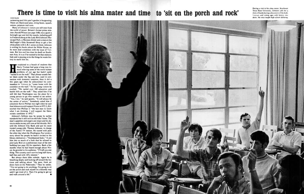 Eine Person, die in einem Raum mit vielen sitzenden Personen, Vorhängen und Fenstern steht und eine Brille trägt.