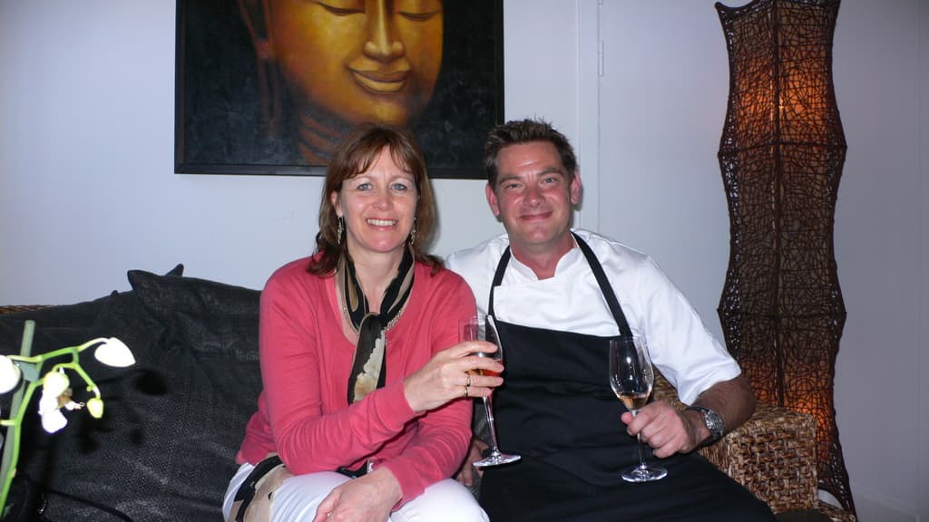 Zwei Personen sitzen auf einer Couch, lächeln und halten Weingläser, mit einer Wand und einem Foto im Hintergrund.