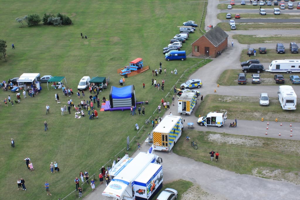 Eine Vogelperspektive auf einen Parkplatz mit vielen Fahrzeugen, ein paar Menschen in der Nähe von Zelten, einer zentralen Hütte und ein paar Bäumen auf der linken Seite.