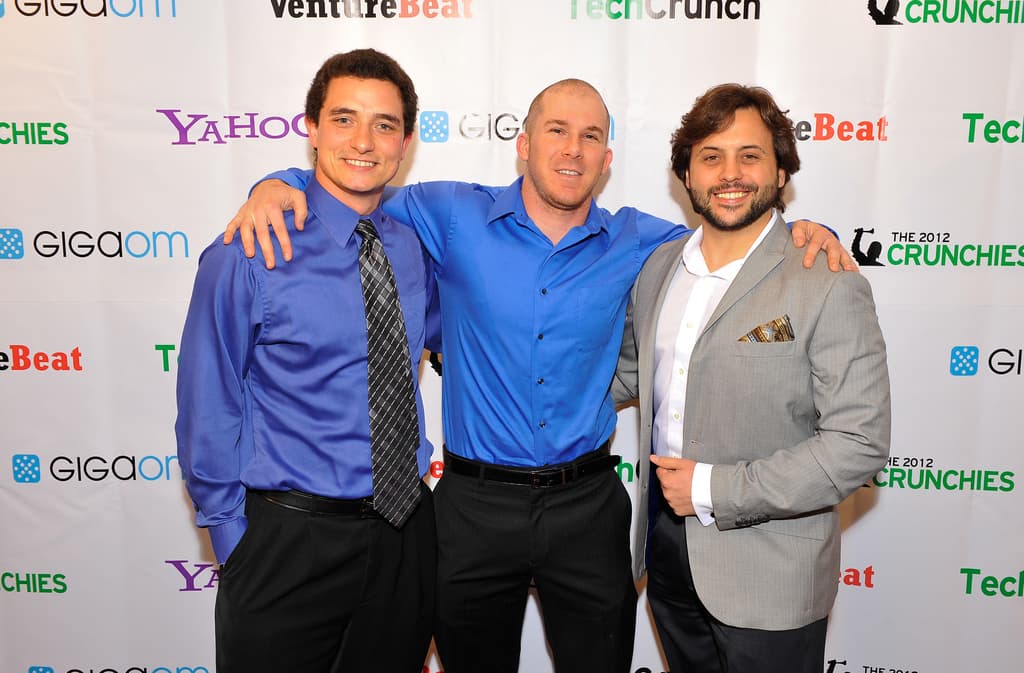 Drei Personen vor einem Sponsorenbanner, alle mit Kleidung.