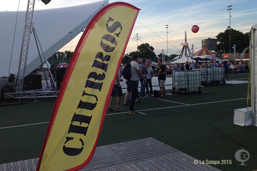 Eine belebte Karnevalsszene mit Zelten, einem zentralen Mast, Menschen, Bäumen, Strommasten, Straßenbahnen auf der Straße und einem eisernen Stand auf der linken Seite unter einem klaren Himmel.