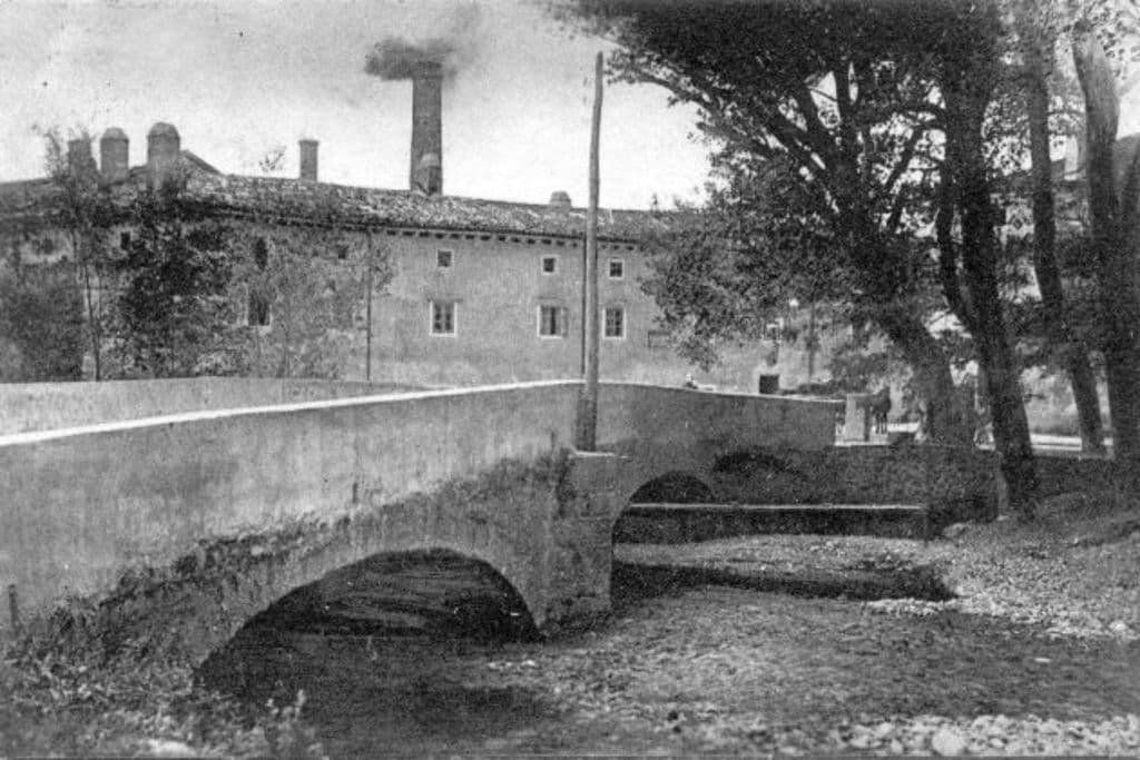 Eine Stadtansicht mit Gebäuden, Bäumen und einer Brücke im Vordergrund, Rauch der aus einem Schornstein aufsteigt, unter einem Himmel mit Wasser am Boden.