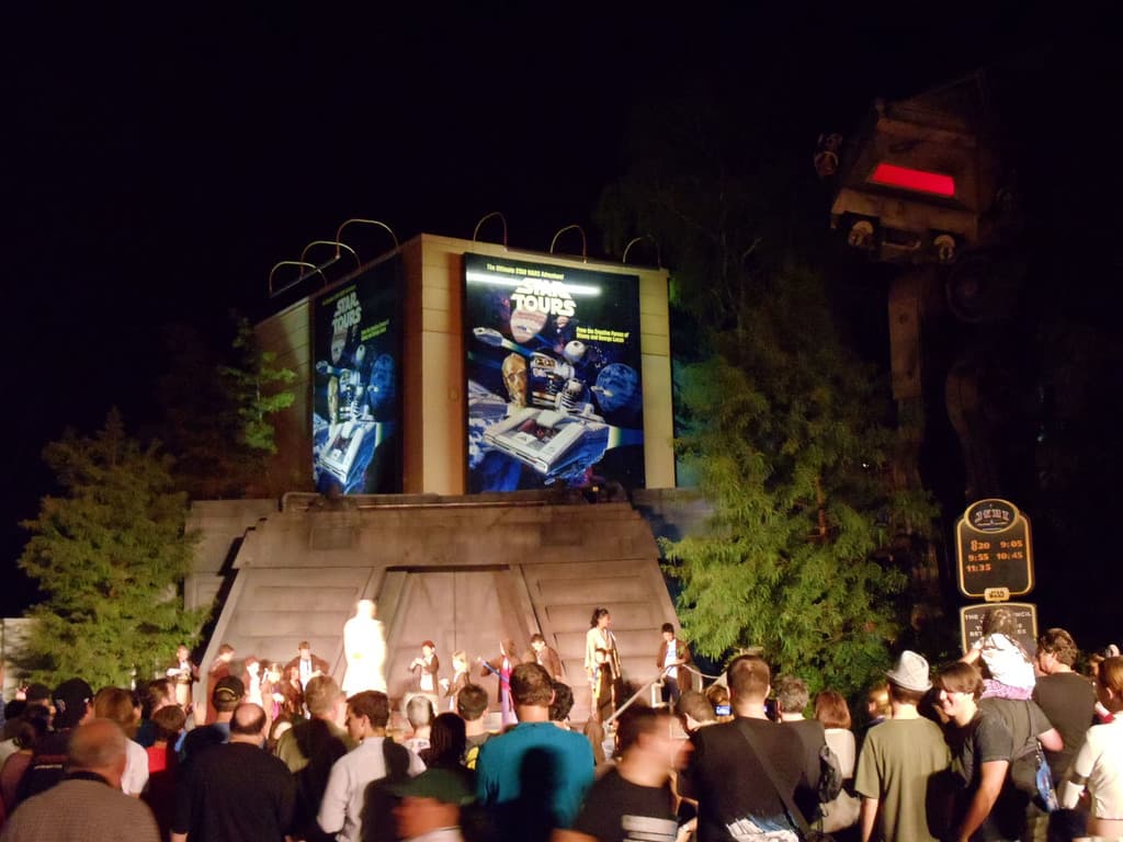 Eine nächtliche Außenansicht mit Menschen unten, Kindern auf der Bühne vor ihnen, einer braunen Wand hinter der Bühne, einem Gebäude mit Werbetafeln dahinter, Bäumen auf beiden Seiten des Gebäudes und Tafeln mit Text auf der rechten Seite.
