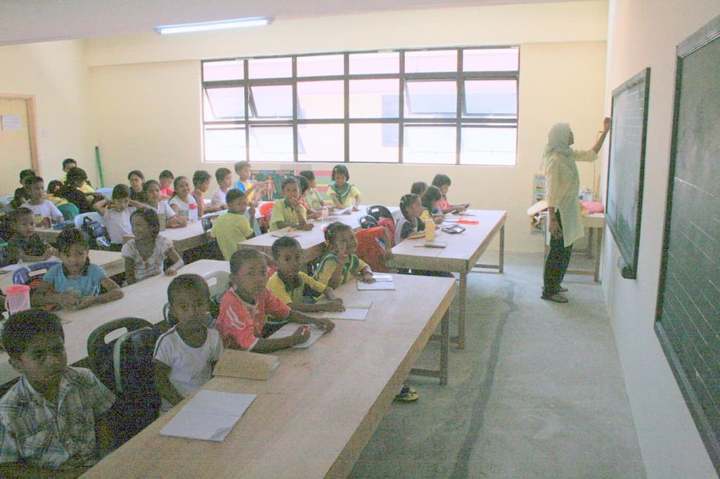 Ein Klassenzimmer mit einer Tafel auf der rechten Seite, an der eine Frau schreibt, Fenster in der Mitte, eine Lampe oben und Tischen mit Stühlen, Büchern und Wasserflaschen, auf denen Kinder sitzen.