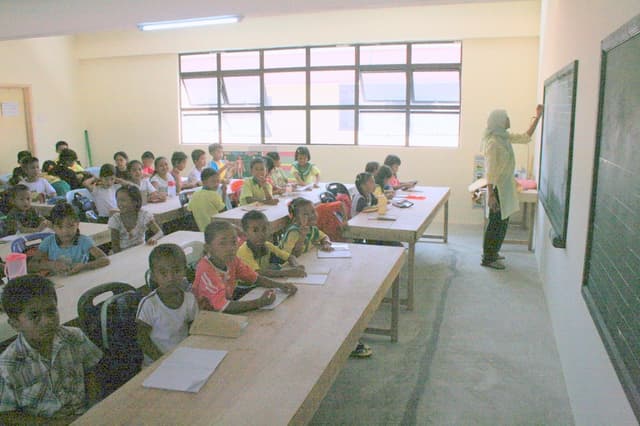 Ein Klassenzimmer mit einer Tafel auf der rechten Seite, an der eine Frau schreibt, Fenster in der Mitte, eine Lampe oben und Tischen mit Stühlen, Büchern und Wasserflaschen, auf denen Kinder sitzen.