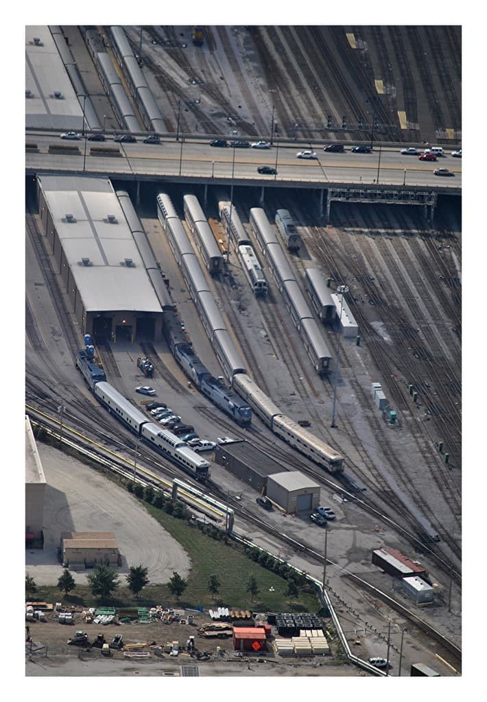 Eine Brücke mit wenigen Fahrzeugen darauf, darunter Eisenbahnschienen mit ein paar Zügen und eine Landschaft mit Gebäuden, Bäumen, Pfählen und diversen Objekten und Fahrzeugen am Boden.