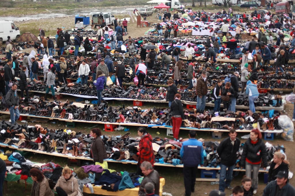 Ein belebtes Marktbild mit zahlreichen Menschen, Tischen mit Schuhen, Kleidung und anderen Gegenständen, einer Tasche rechts und Fahrzeugen und Zelten im Hintergrund.