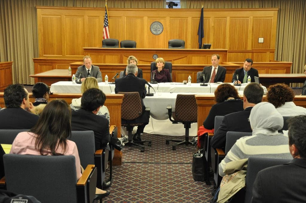 Ein Versammlungsraum mit sitzenden Menschen, zwei ausländischen Flaggen, einem orangen Schreibtisch, Wasserflaschen und einem Mikrofon.