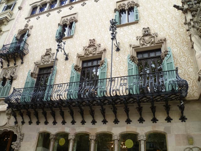 Ein Gebäude mit Fenstern, Balkonen mit Stangen und Wandreliefs, mit einem Kronleuchter im Inneren am unteren Bildrand sichtbar.
