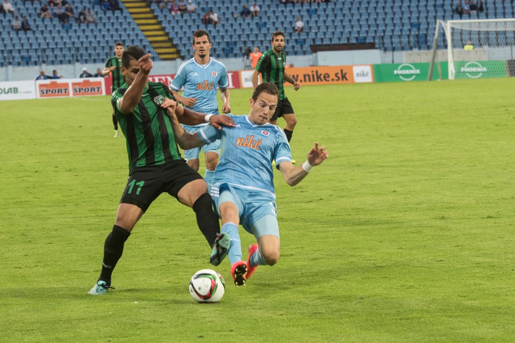 Menschen spielen ein Spiel auf einem grünen Feld mit einem Ball, Netz und Werbetafeln, während das Publikum von Stadionstühlen im Hintergrund zuschaut.