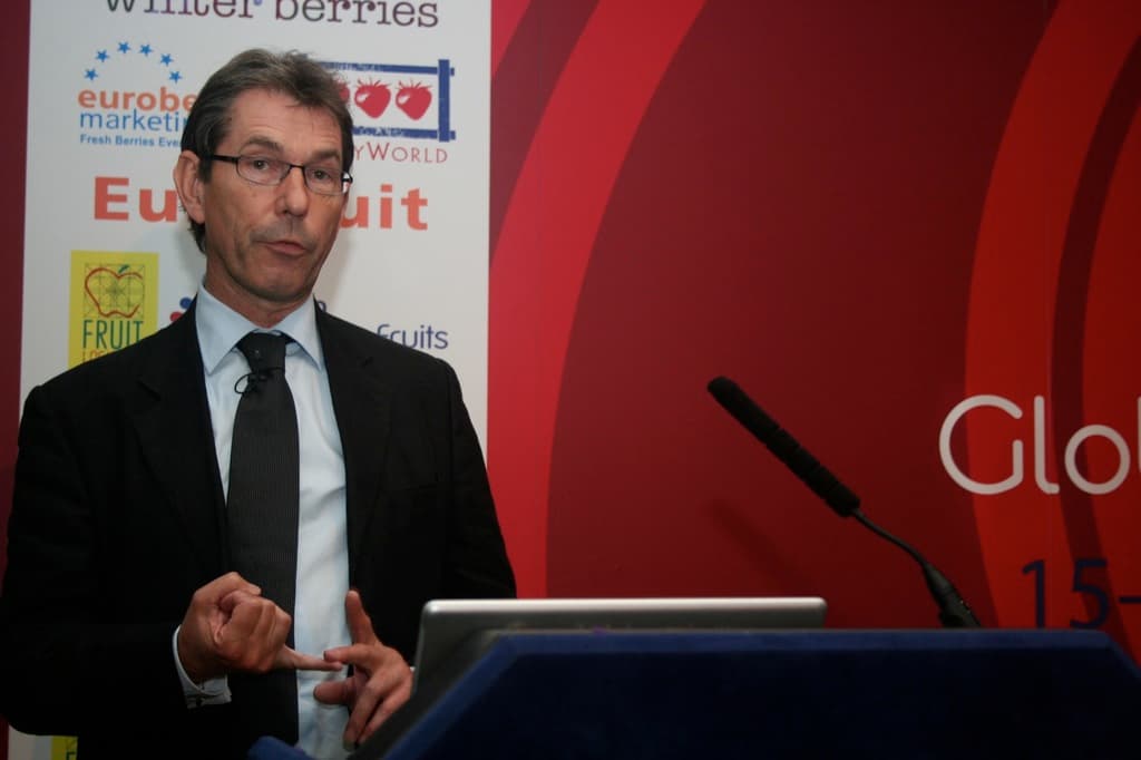 Ein Mann steht an einem Podium mit einem Mikrofon und Laptop, vor einer roten Fahne mit Text.