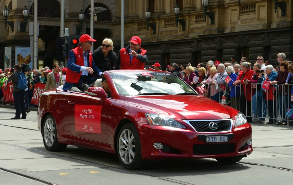 Ein Auto mit Menschen darin fährt auf einer Straße, mit verschiedenen Objekten, Menschen und Strukturen im Hintergrund.