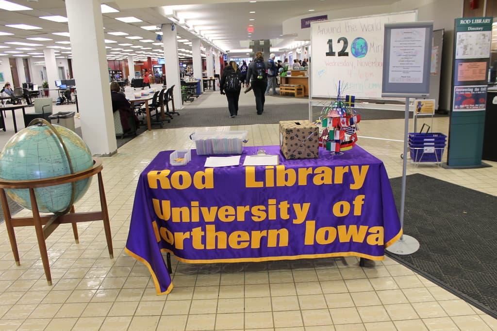 Eine Ausstellung mit einem Tisch, auf dem verschiedene Landesflaggen, eine Box, Papiere und ein Banner stehen, eine Kugel zur Linken, eine Mitteilungstafel zur Rechten mit einigen Menschen in der Nähe und mehreren Tischen und Stühlen zur Linken.