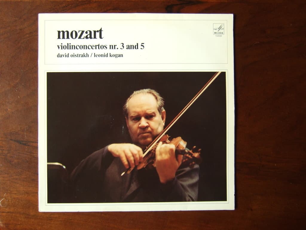 Ein Mann in einem schwarzen Mantel spielt Geige auf einem Holztisch, mit schwarzem Hintergrund und einem kleinen Zitat oben auf dem Bild.