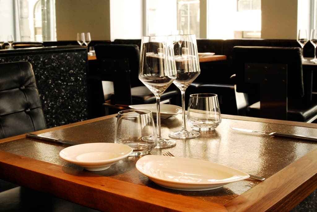 Ein Restaurantinnenraum mit einem Tisch, der mit Gläsern und Tellern gedeckt ist, umgeben von Stühlen und einer Wand mit Fenstern im Hintergrund.