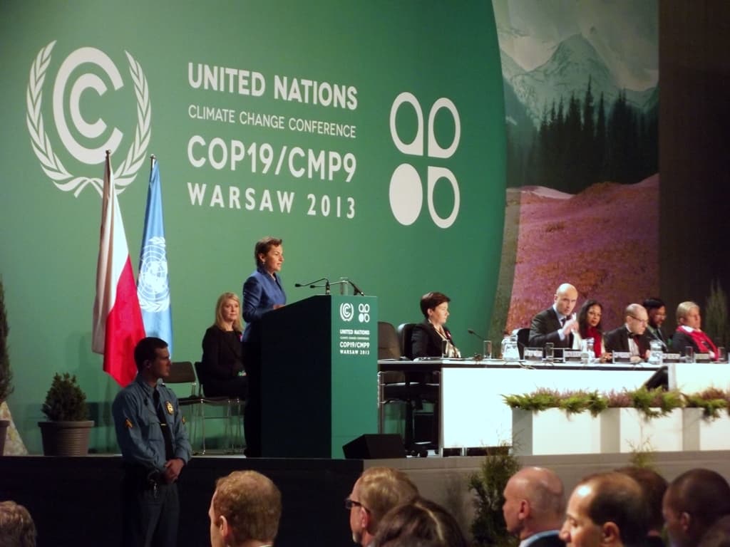 Eine Frau steht neben einem Podium mit Mikrofonen und spricht zu einem sitzenden Publikum. Ein Tisch mit Gläsern, Mikrofonen, Namensschildern und anderen Gegenständen ist im Vordergrund zu sehen. Zwei Flaggen hängen an Stangen, und ein Blumentopf mit einer Pflanze ist sichtbar. Kleine Pflanzen und eine Werbetafel sind ebenfalls vorhanden. Eine weitere Person steht in der Nähe, und eine Gruppe von Menschen ist unten im Bild zu sehen.