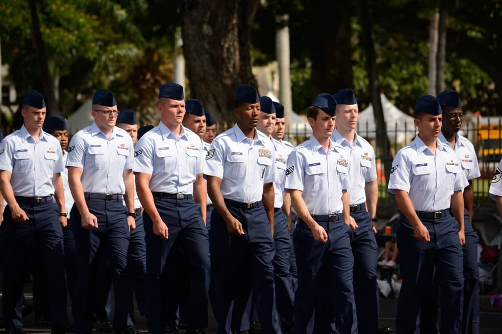 Männer in Uniform gehen auf einer Straße mit einem Eisenzaun dahinter, Bäume und Pfosten im Hintergrund.