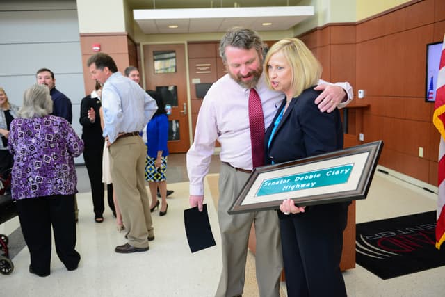 Eine Gruppe von Menschen, die drinnen stehen, wobei einer ein Foto mit 'Senator Debbie Clary Highway' darauf hält, mit einer Flagge, einer Wand, einer Tür und Lichtern im Hintergrund.