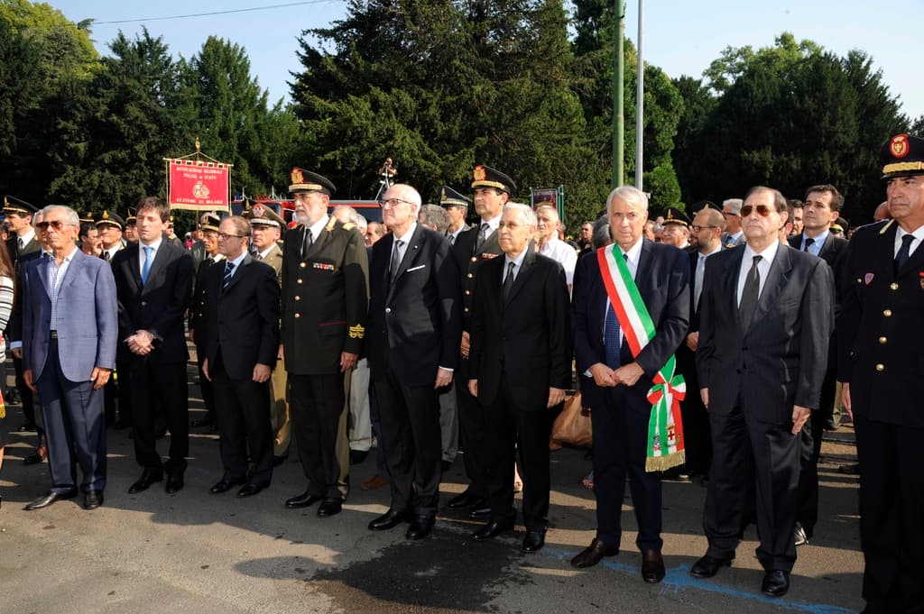 Mehrere Personen in Anzügen stehen im Freien mit zwei Pfählen und Bäumen im Hintergrund.