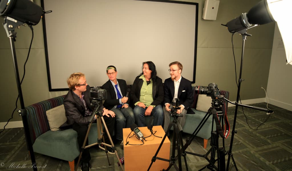 Eine Gruppe von drei Personen sitzt auf einem Sofa und gibt ein Interview, wobei eine Person in einem schwarzen Mantel und Hose links, eine Person in einem Mantel und blauer Krawatte mit einem Mikrofon in der Hand in der Mitte und eine Person rechts mit einem Lächeln zu sehen ist. Hinter ihnen befindet sich ein Projektionsschirm, ein Mikrofonrekorder auf dem Tisch in der Mitte und Kameras mit großen Stativen auf beiden Seiten.