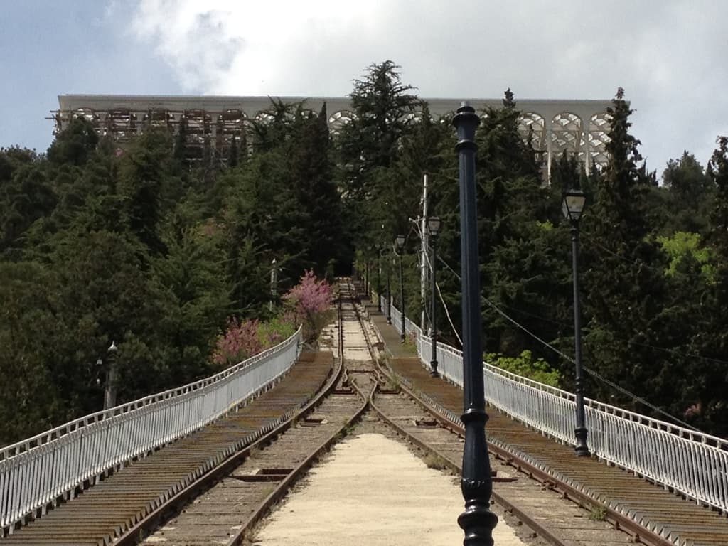 Eisenbahnschienen mit einem mittigen Pfahl und Bäumen, flankiert von Pfählen und Zaun, mit einem Gebäude und Himmel im Hintergrund.