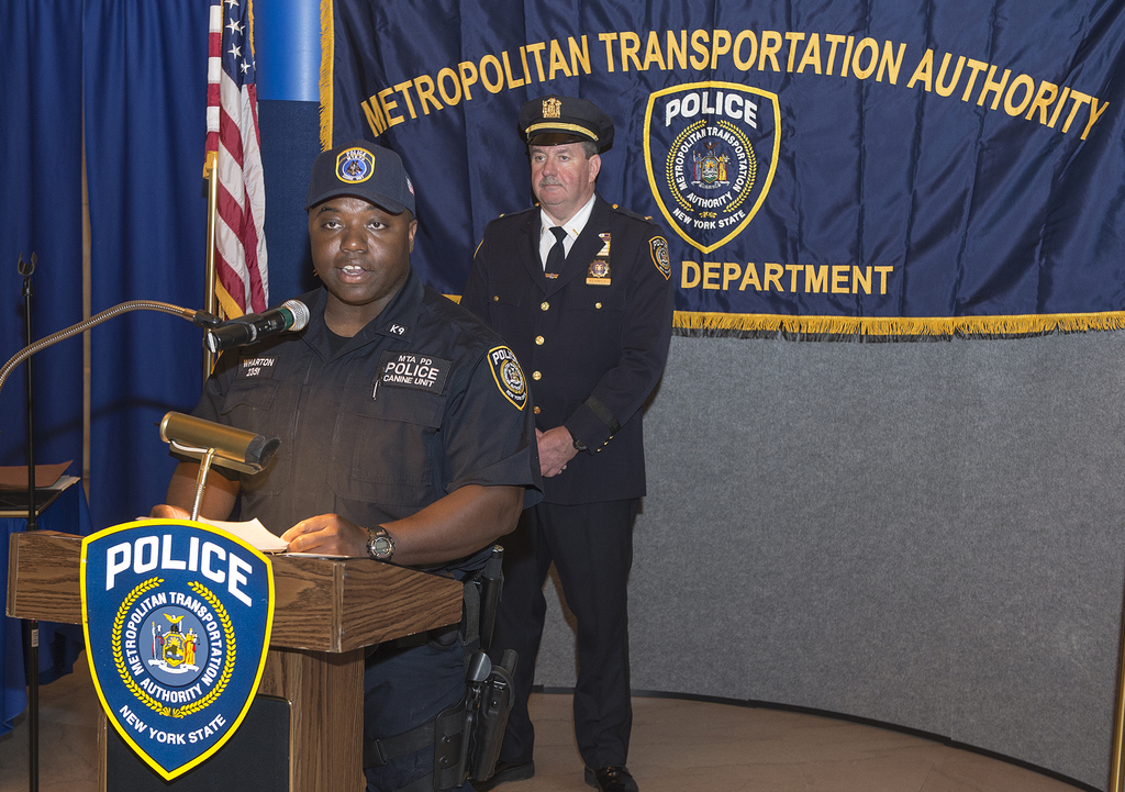 Zwei Polizeibeamte stehen vor einem Rednerpult, einer hält Papiere und spricht in ein Mikrofon, mit einer Fahne, einem Banner und einem Tisch in der Nähe, und einer Tafel mit Symbolen und Text im Hintergrund.