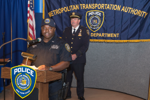 Zwei Polizeibeamte stehen vor einem Rednerpult, einer hält Papiere und spricht in ein Mikrofon, mit einer Fahne, einem Banner und einem Tisch in der Nähe, und einer Tafel mit Symbolen und Text im Hintergrund.